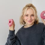 Close up portrait of happy, cute blond woman, holding doughnut, eating sweet, delicious comfort food, showing dessert at camera